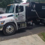 A white dump truck parked on a residential street with contact information and American flag decals on the side.