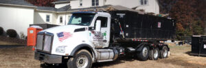 A white semi-truck with an attached black dumpster parked on dry grass near a white building and portable toilets.