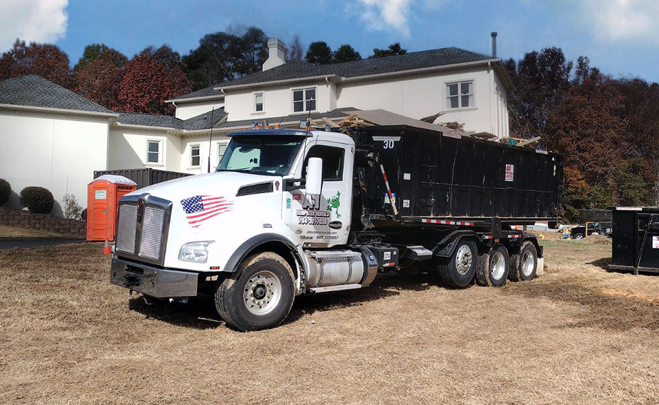 A white semi-truck with an attached black dumpster parked on dry grass near a white building and portable toilets.