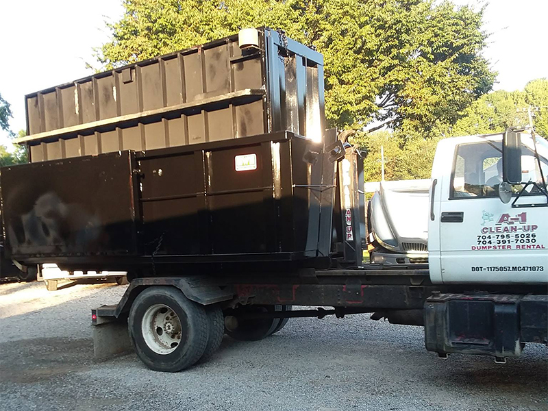 A black dumpster on a white truck parked on a gravel surface with trees in the background.