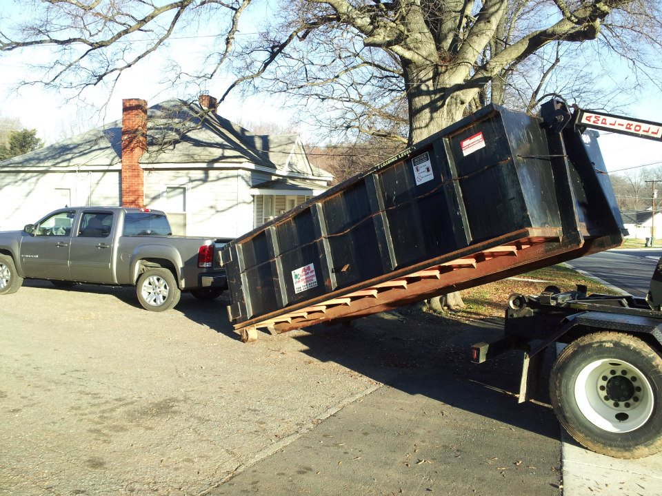 A large black dumpster is being loaded onto a tow truck next to a gray pickup truck near a house with a brick chimney.