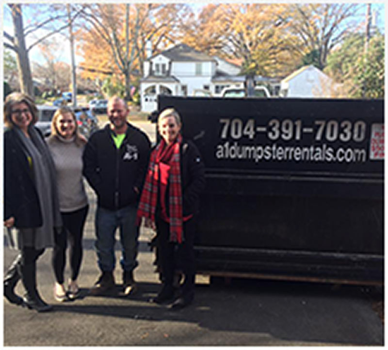 Four people stand in front of a large black dumpster outside, with autumn trees and houses in the background.